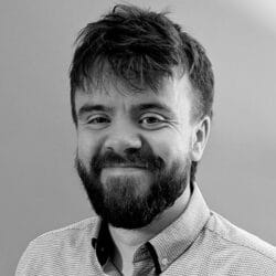 Sam Ward, a bearded man with short, tousled hair, smiles slightly while wearing a light-colored, patterned button-up shirt against a plain background.