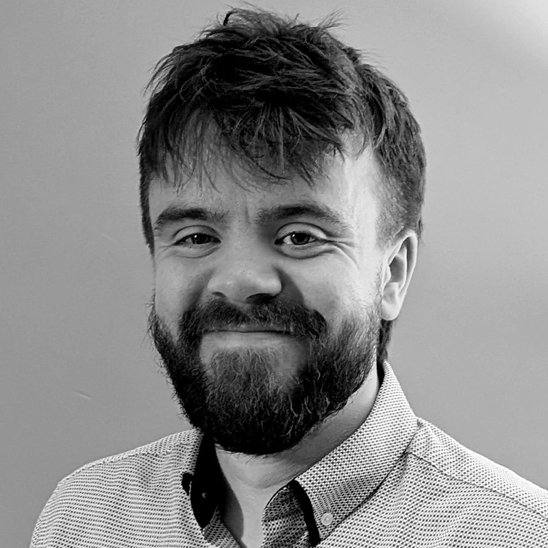 Sam Ward, a bearded man with short, tousled hair, smiles slightly while wearing a light-colored, patterned button-up shirt against a plain background.