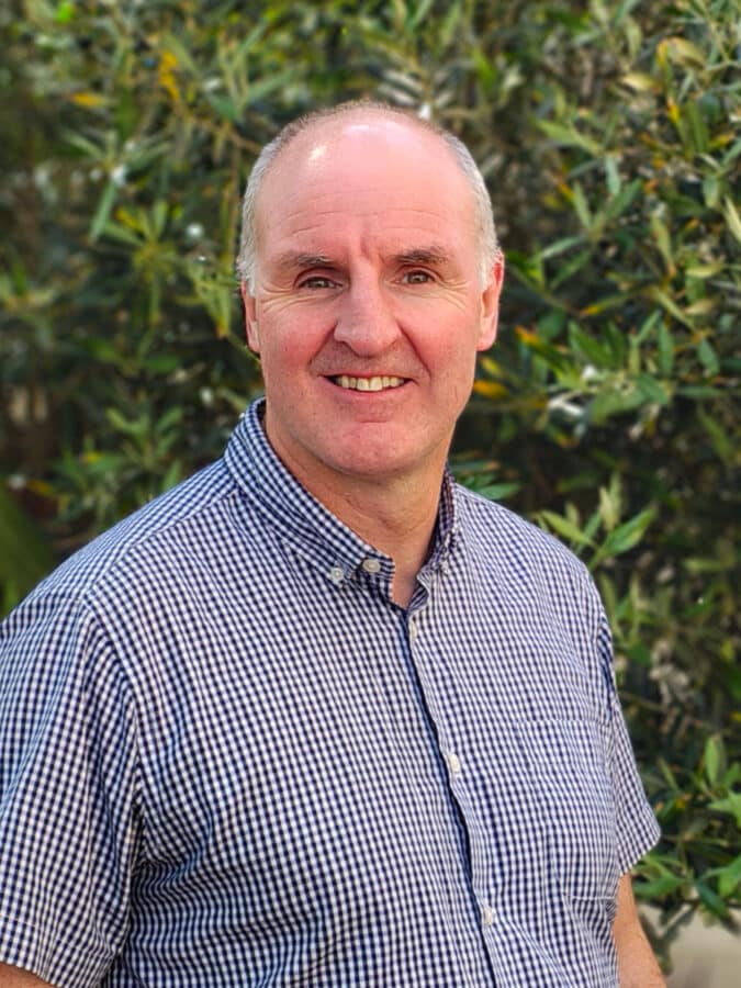 Barrie Anderson, consultant at Create Consulting Engineers, smiling outdoors wearing a blue checked shirt against a green plant background.