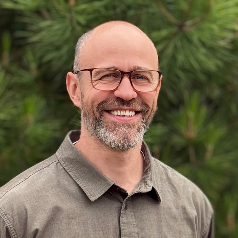 Ben Dixon, Principal Acoustics Consultant at Create Consulting Engineers, smiling outdoors against a green foliage background.