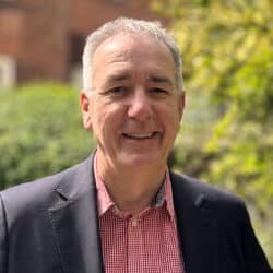 Jonathan Cage, consultant at Create Consulting Engineers, smiling outdoors in professional attire with brick building and greenery background.