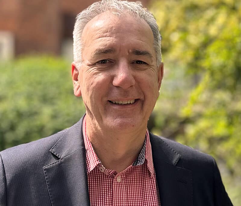 Jonathan Cage, consultant at Create Consulting Engineers, smiling outdoors in professional attire with brick building and greenery background.