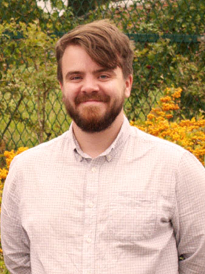 Sam Ward, consulting engineer at Create Consulting Engineers, wearing a light beige shirt against a garden background with ivy and yellow flowers.