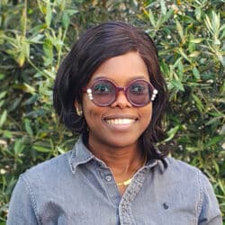 Professional headshot of Bomi Ogunade at Create Consulting Engineers, smiling against a green foliage background, wearing glasses and denim shirt.