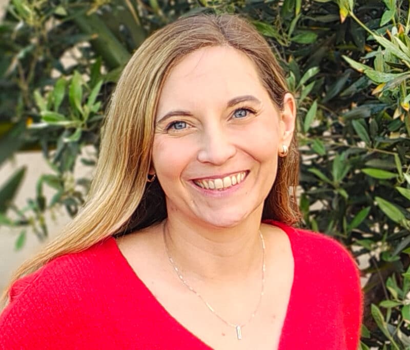 Professional headshot of a woman wearing a red top, smiling outdoors against green foliage backdrop.