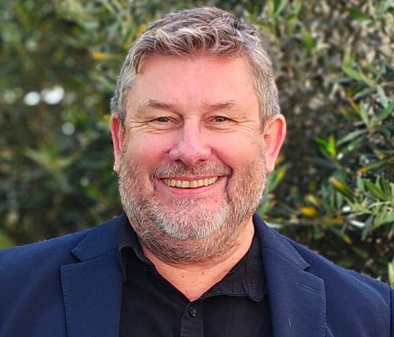 Professional headshot of a man with grey hair and beard wearing a dark navy blazer and black shirt, photographed outdoors with green foliage background.