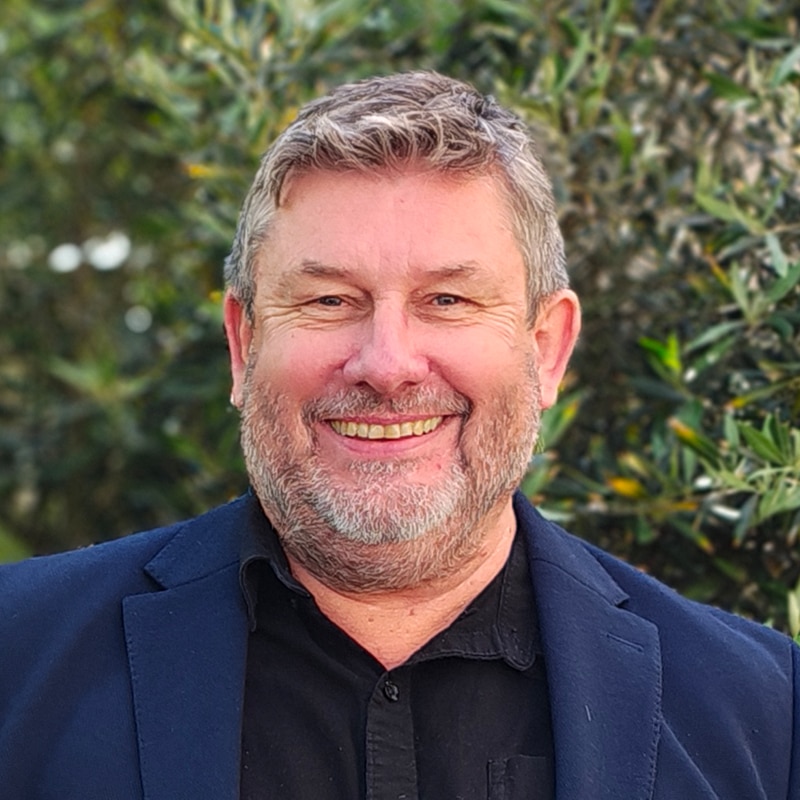 Professional headshot of a man with grey hair and beard wearing a dark navy blazer and black shirt, photographed outdoors with green foliage background.