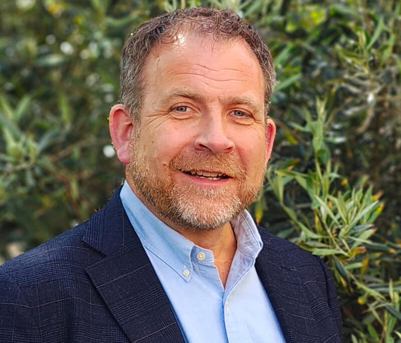 Professional portrait of a consulting engineer at Create Consulting Engineers, smiling outdoors with foliage background, wearing navy blazer and light blue shirt.