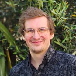 Professional portrait of a male consultant wearing glasses and patterned dark shirt against green foliage background.