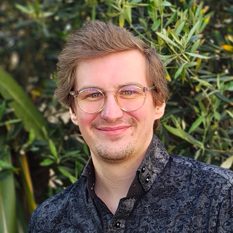 Professional portrait of a male consultant wearing glasses and patterned dark shirt against green foliage background.