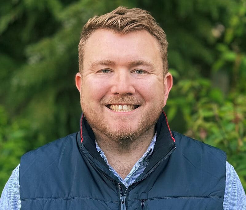 Professional headshot of a male engineer smiling outdoors with green foliage background, wearing navy gilet and blue shirt.