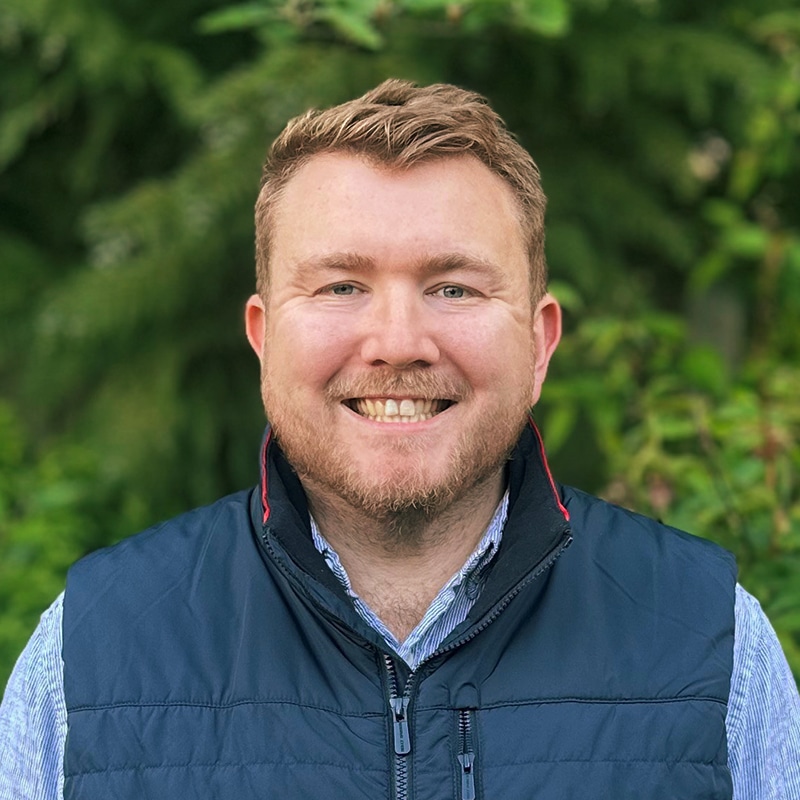 Professional headshot of a male engineer smiling outdoors with green foliage background, wearing navy gilet and blue shirt.