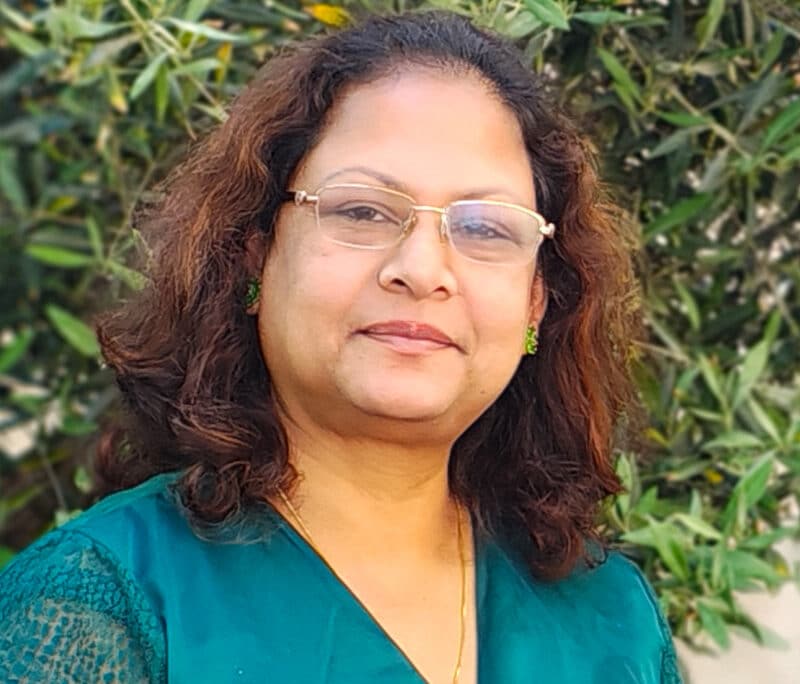 Professional portrait of a woman wearing glasses and teal top against green foliage background
