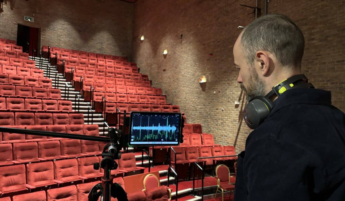 A man wearing headphones monitors acoustics and audio levels on a tablet in an empty theater with red seats.
