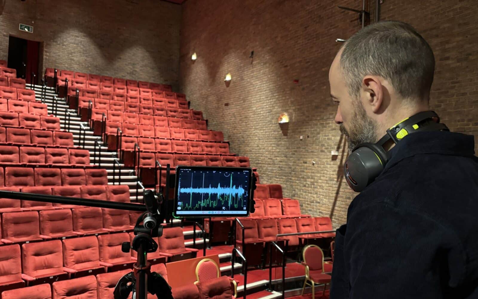 A man wearing headphones monitors acoustics and audio levels on a tablet in an empty theater with red seats.