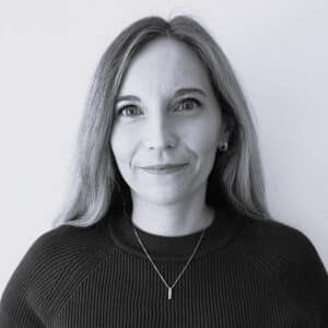 Claire Burroughs, a woman with long, straight hair wearing a dark sweater and a necklace, stands against a plain light background, looking at the camera with a neutral expression.