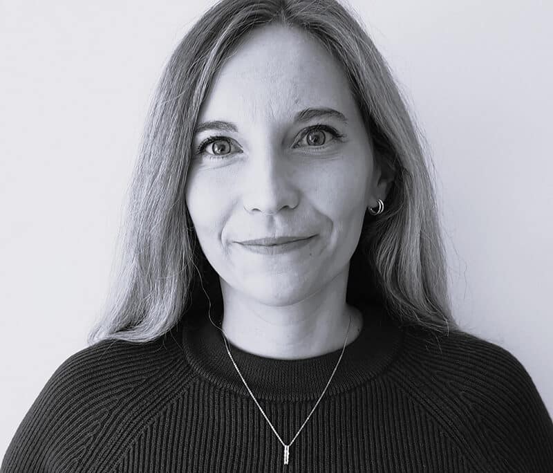 Claire Burroughs, a woman with long, straight hair wearing a dark sweater and a necklace, stands against a plain light background, looking at the camera with a neutral expression.