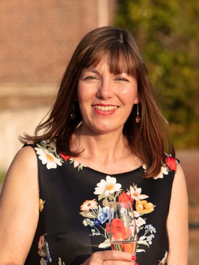 Karen, half-body portrait, smiling whilst holding a glass of water, wearing a black floral sleeveless top outdoors.