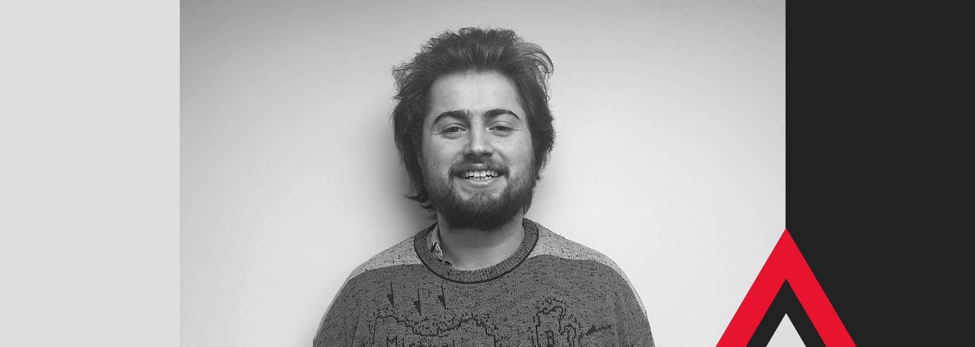 George Baker, a person with short, dark hair and a beard, smiles at the camera, standing against a plain light background with abstract blue, black, and red geometric shapes framing the image.