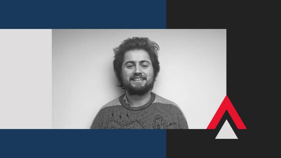 George Baker, a person with short, dark hair and a beard, smiles at the camera, standing against a plain light background with abstract blue, black, and red geometric shapes framing the image.