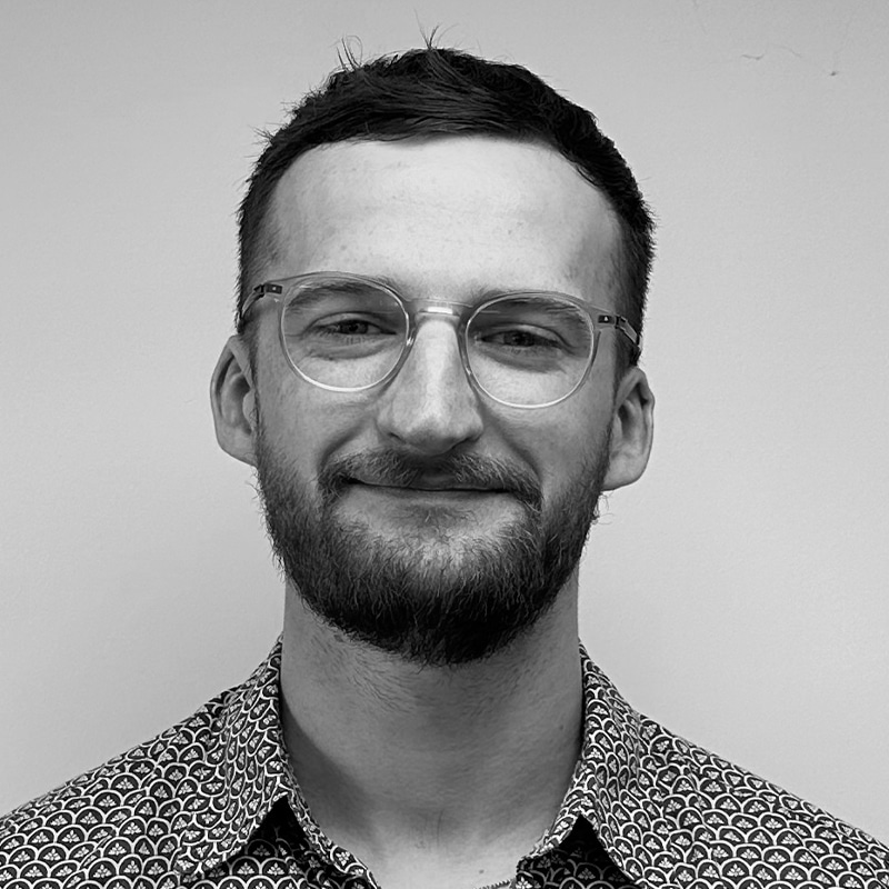 Martyn Broom, a man with short dark hair, glasses, and a beard, smiles slightly at the camera. He is wearing a patterned shirt and is posed against a plain light-colored background.