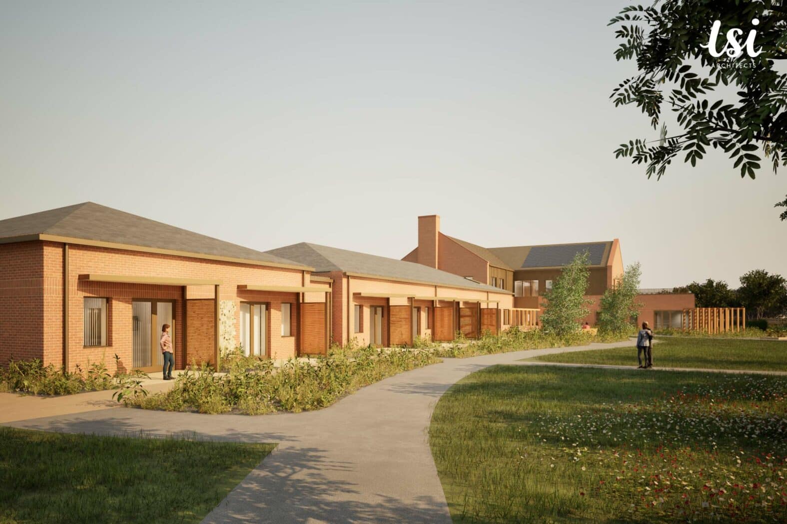 A paved path curves through a grassy area in front of a row of single-story brick buildings with covered porches; two people are visible outside.