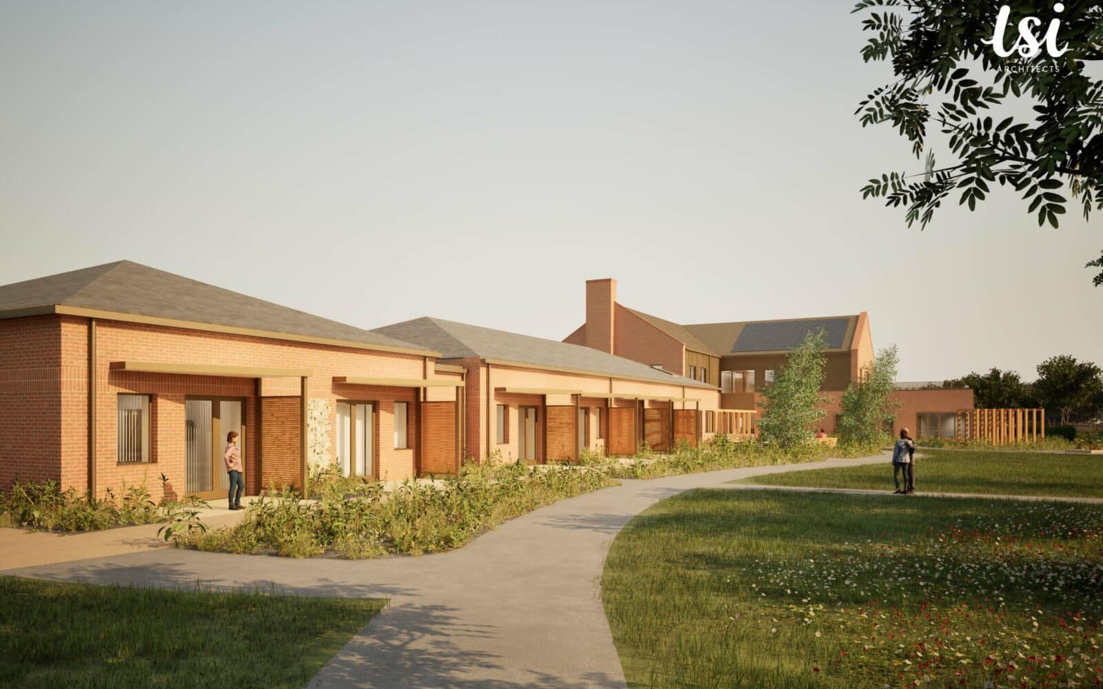 A paved path curves through a grassy area in front of a row of single-story brick buildings with covered porches; two people are visible outside.
