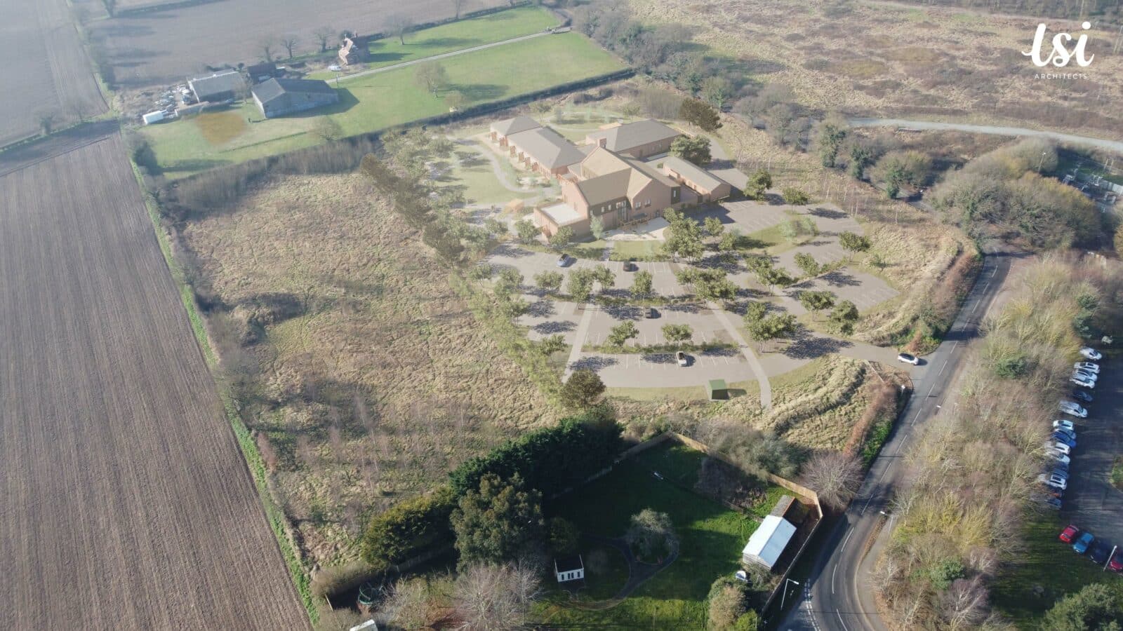 Aerial view of a large building complex surrounded by fields, trees, and roads, with parking lots and several vehicles visible.