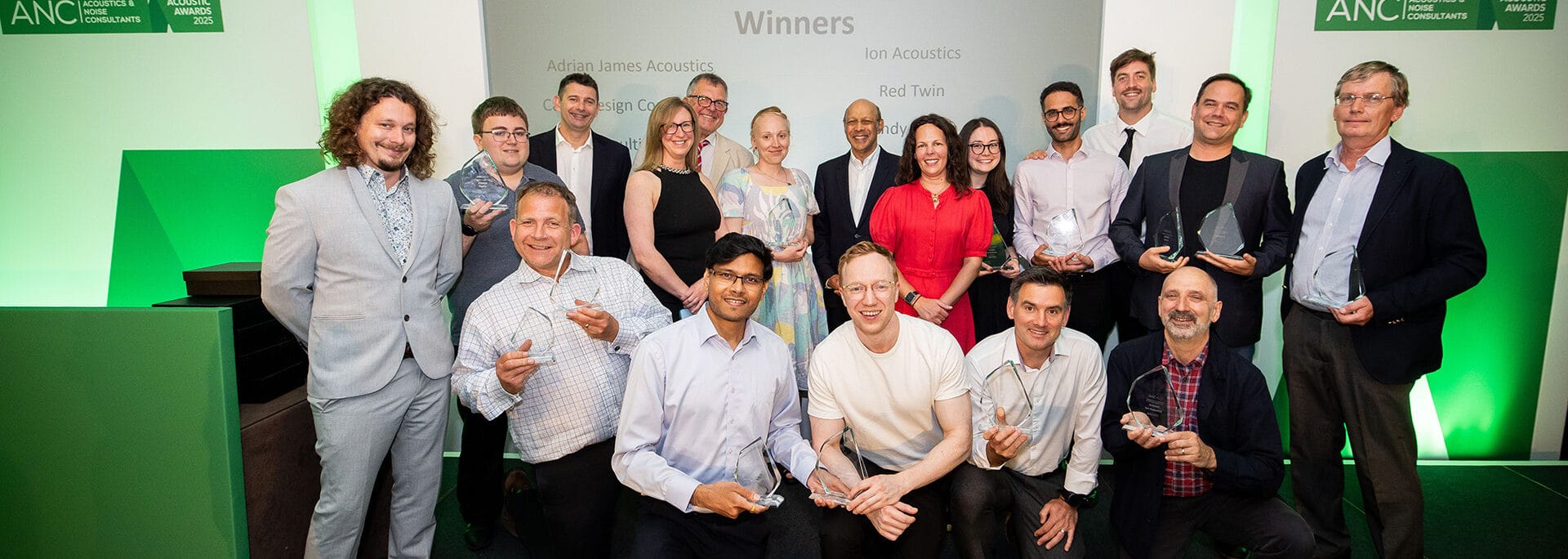 A group of people pose on stage holding trophies at an awards event in front of a screen displaying the word "Winners.