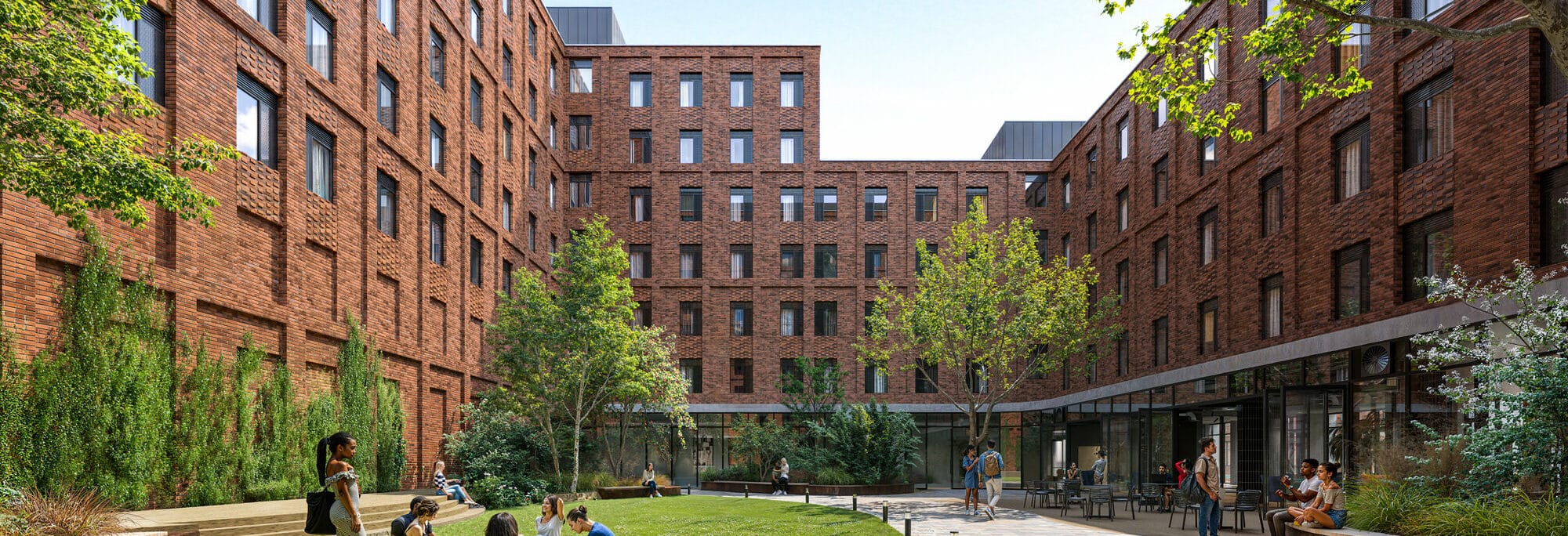 A group of people sit and stand on the grass and pathways in a courtyard surrounded by multi-story brick buildings, with trees and shrubs scattered around.