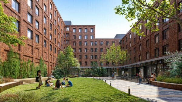 A group of people sit and stand on the grass and pathways in a courtyard surrounded by multi-story brick buildings, with trees and shrubs scattered around.