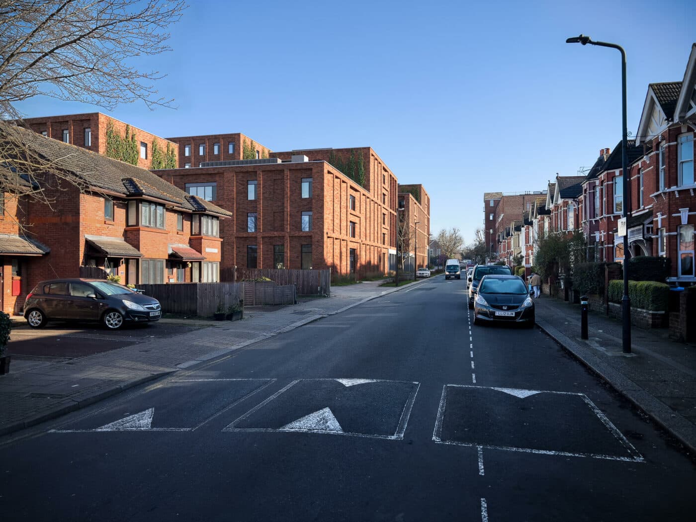 A quiet residential street with parked cars, brick houses on the left, terraced homes on the right, and speed bumps on the road under a clear blue sky.