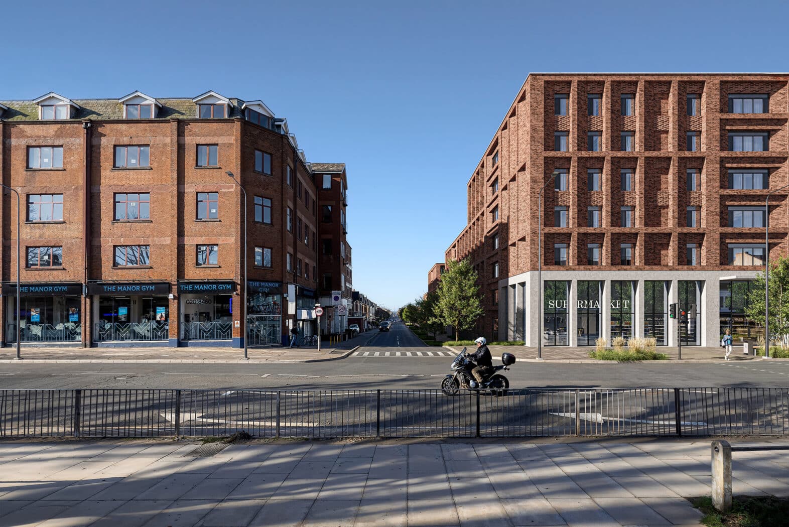 A person on a motorcycle rides across an intersection between two modern brick buildings under a clear blue sky.
