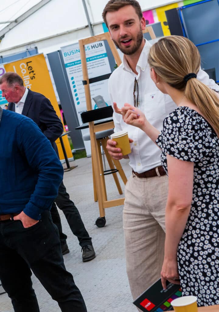 Several people stand and talk at an indoor event or exhibition. A man in a white shirt holds a drink cup, while a woman in a patterned dress gestures as she discusses Careers at Create with him.