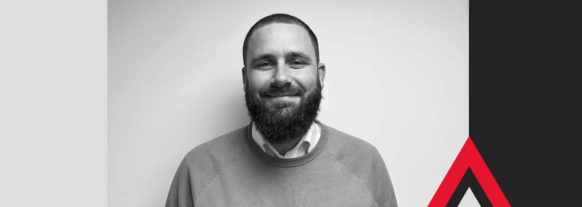 A man with a beard and short hair is smiling at the camera; the image background features geometric shapes in blue, black, white, and red.