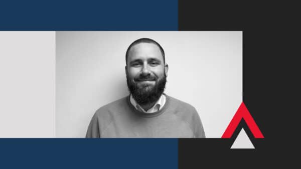 A man with a beard and short hair is smiling at the camera; the image background features geometric shapes in blue, black, white, and red.