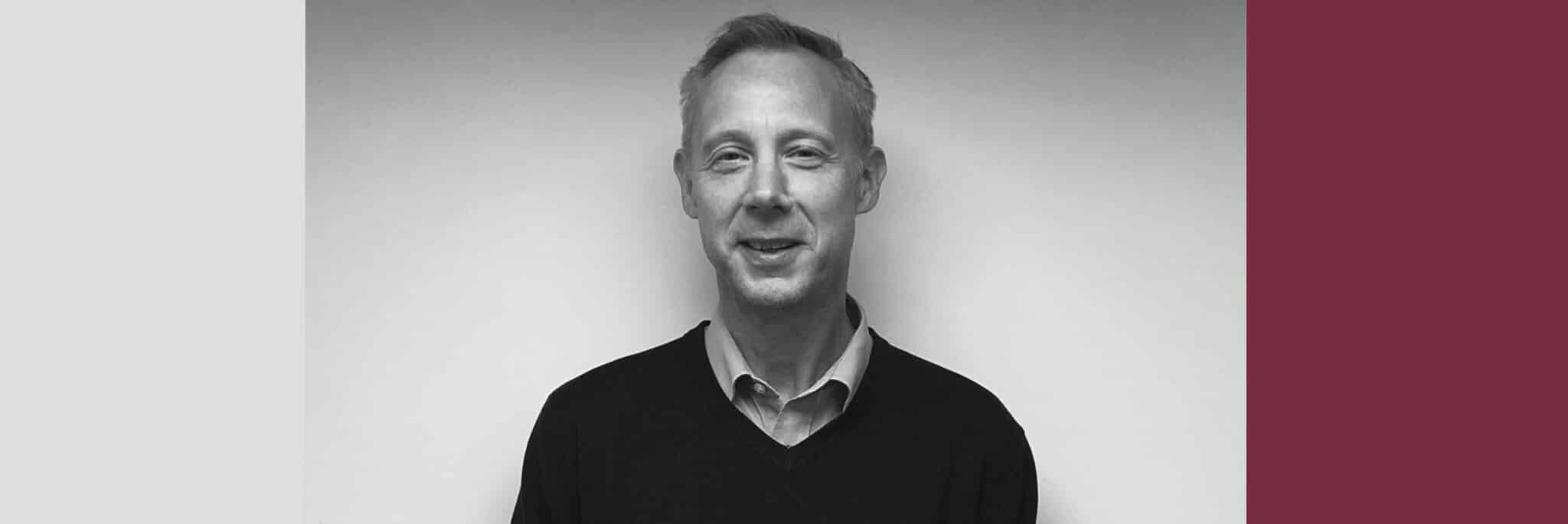 Black and white portrait of a man with short hair wearing a collared shirt and sweater, standing against a plain wall with a color-blocked graphic background.