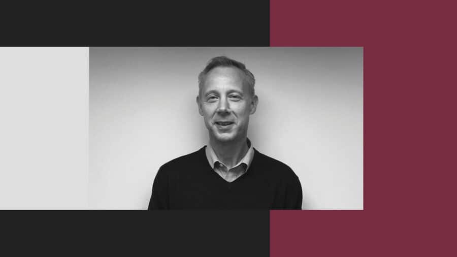 Black and white portrait of a man with short hair wearing a collared shirt and sweater, standing against a plain wall with a color-blocked graphic background.