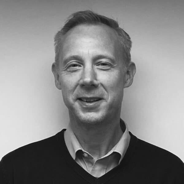 A man with short light hair is smiling, wearing a collared shirt under a dark sweater, standing against a plain light-colored background. The photo is in black and white.