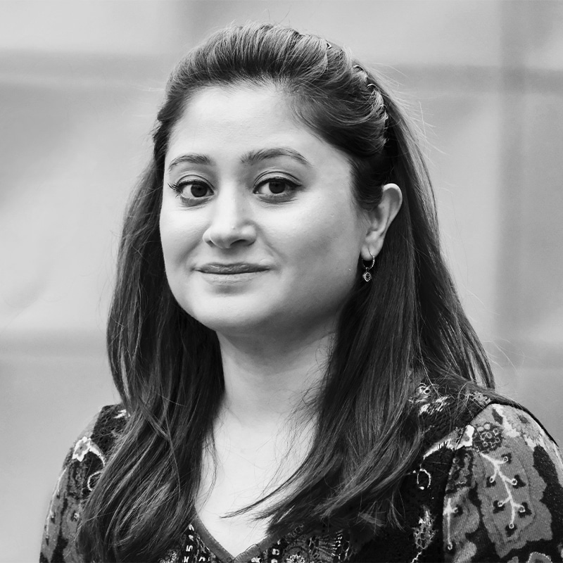 A woman with long, dark hair, wearing a patterned top, looks at the camera with a neutral expression against a blurred background.