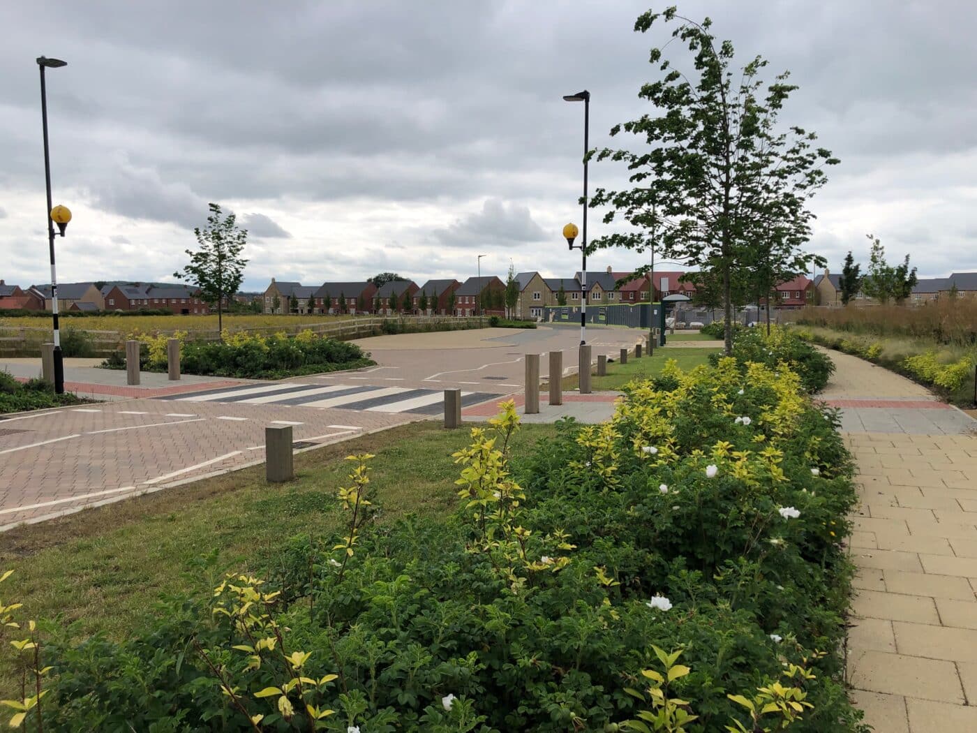 A quiet residential street in Bicester with a crosswalk, streetlights, landscaped bushes, and a row of houses under a cloudy sky.