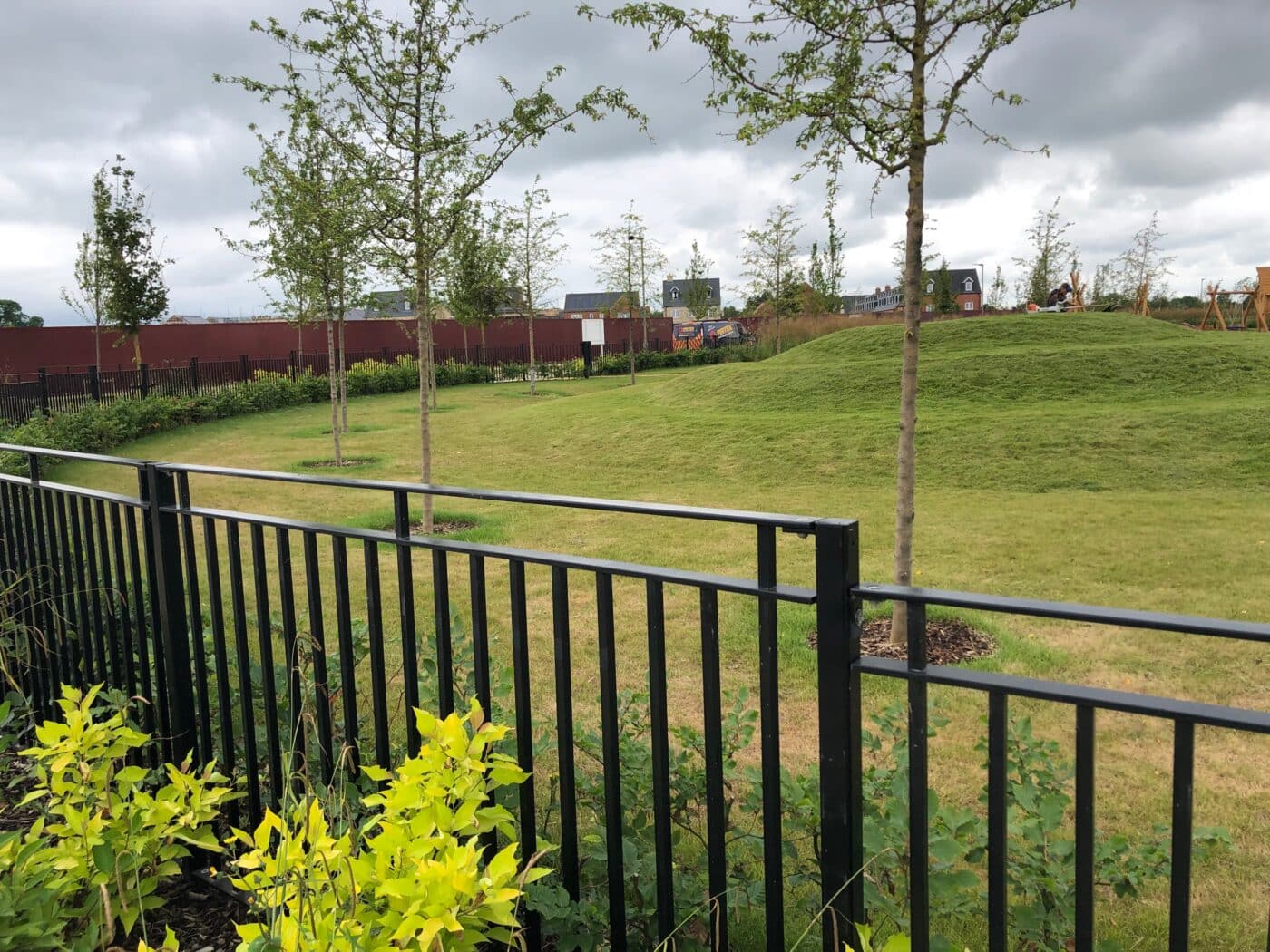 A grassy park area in Bicester with young trees, a black metal fence in the foreground, and cloudy skies above. A red fence and buildings are visible in the background.