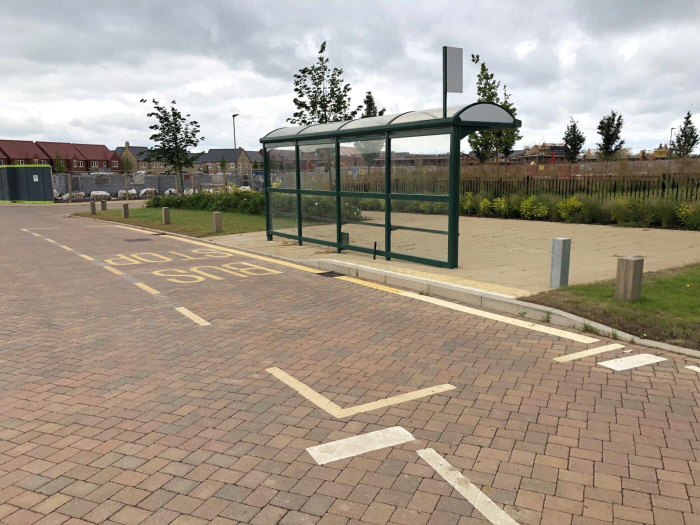 A covered bus stop with a glass shelter is situated on a paved area beside a marked bus stop lane in Bicester, with some trees and buildings in the background under a cloudy sky.