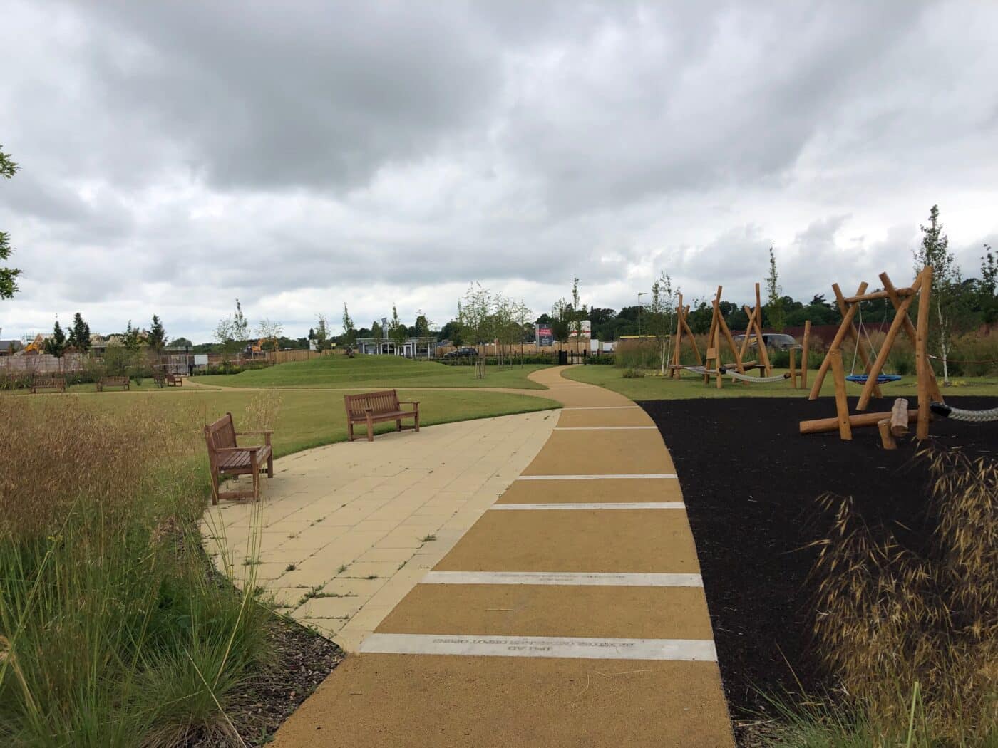 A paved path curves through Bicester park with benches, grassy areas, and a playground featuring wooden swings under a cloudy sky.