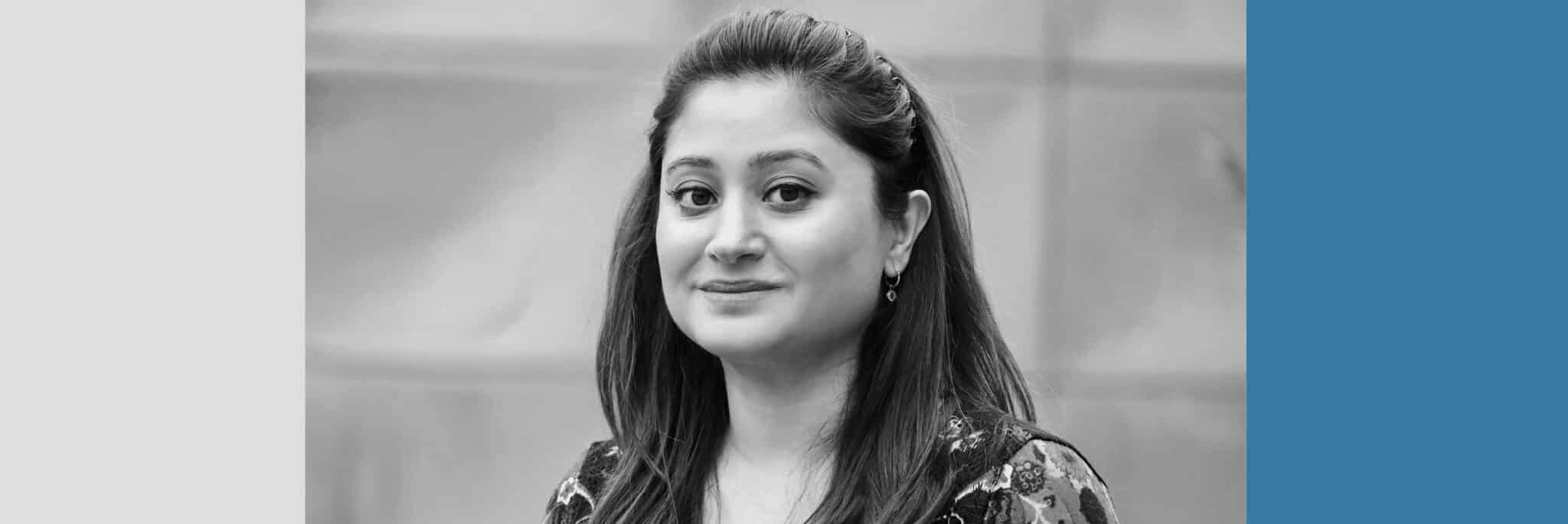 A woman with long dark hair looks at the camera and smiles softly. The photo is black and white, with a gray background and graphic color blocks around the image.