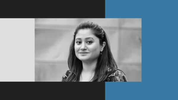 A woman with long dark hair looks at the camera and smiles softly. The photo is black and white, with a gray background and graphic color blocks around the image.