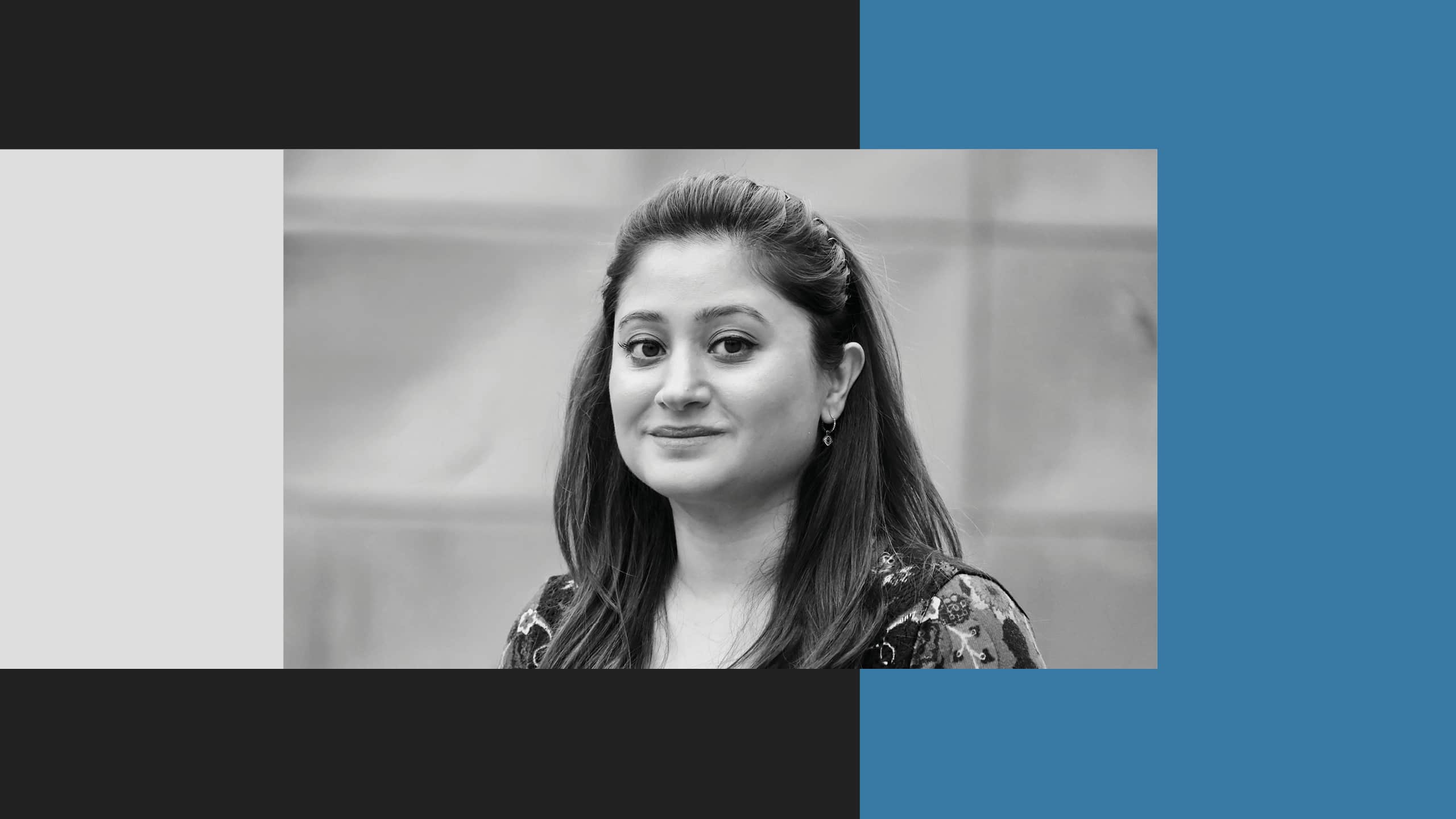 A woman with long dark hair looks at the camera and smiles softly. The photo is black and white, with a gray background and graphic color blocks around the image.