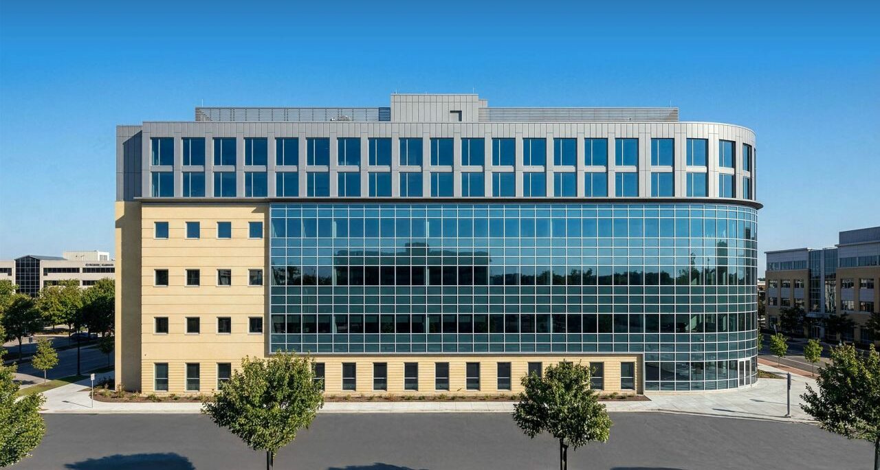 A modern, six-story office building with a curved glass facade and tan stone walls, surrounded by an empty parking lot and small trees.