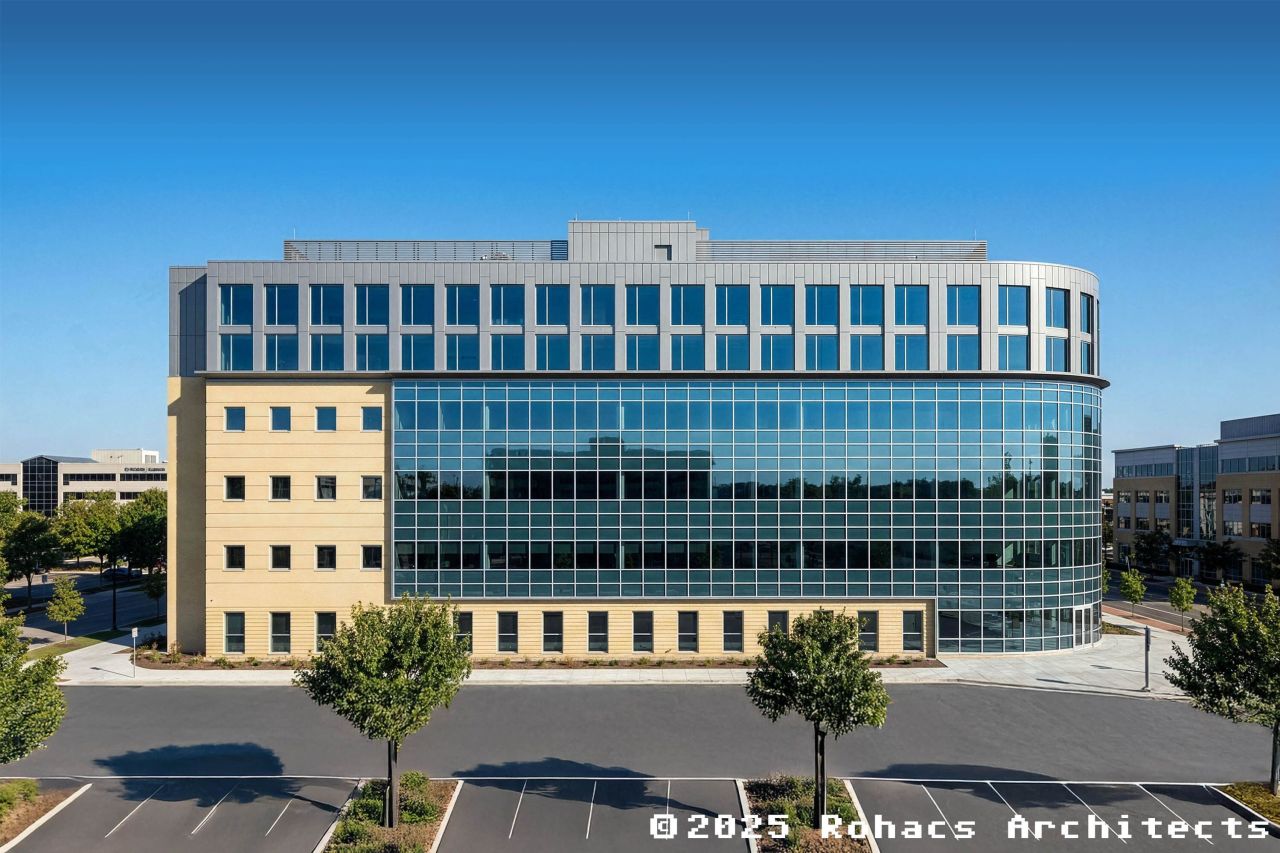 A modern, six-story office building with a curved glass facade and tan stone walls, surrounded by an empty parking lot and small trees.
