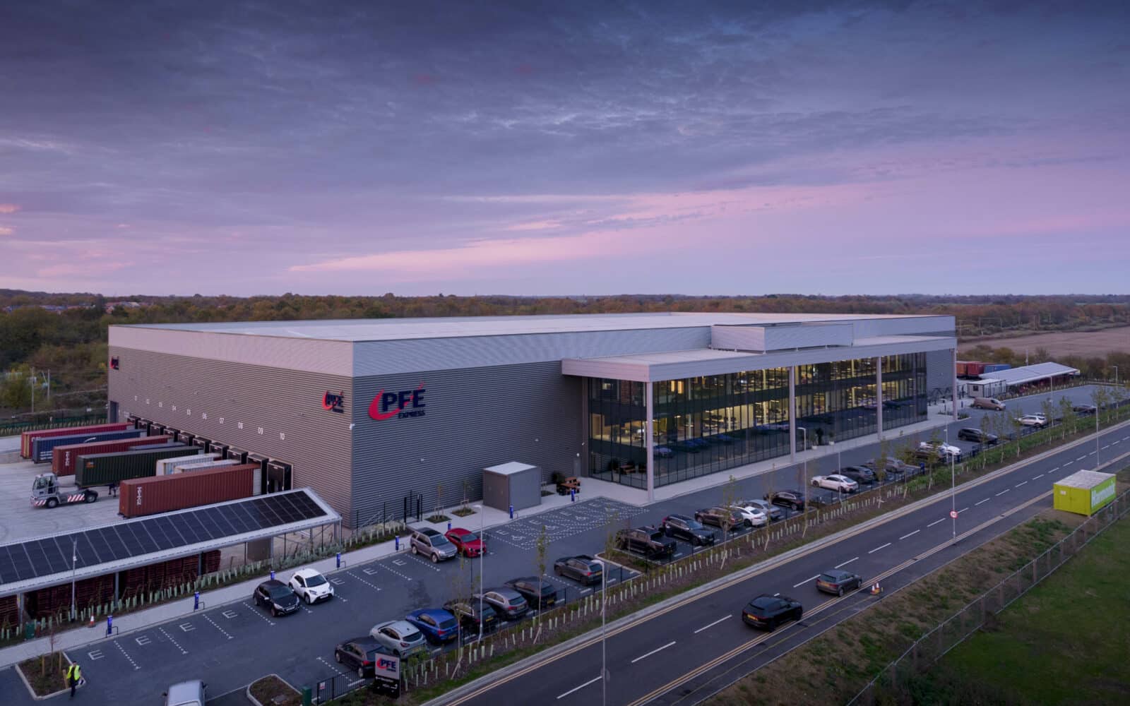 A large warehouse and distribution center with trucks at loading bays, a parking lot with cars, and a road running alongside the building, photographed at dusk.